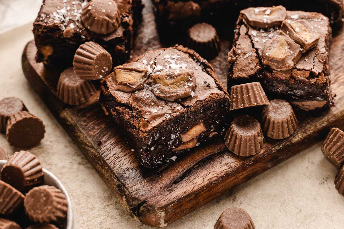 Fudgy chocolate brownies topped with chopped peanut butter cups and a sprinkle of sea salt, displayed on a wooden board with more peanut butter cups scattered around.