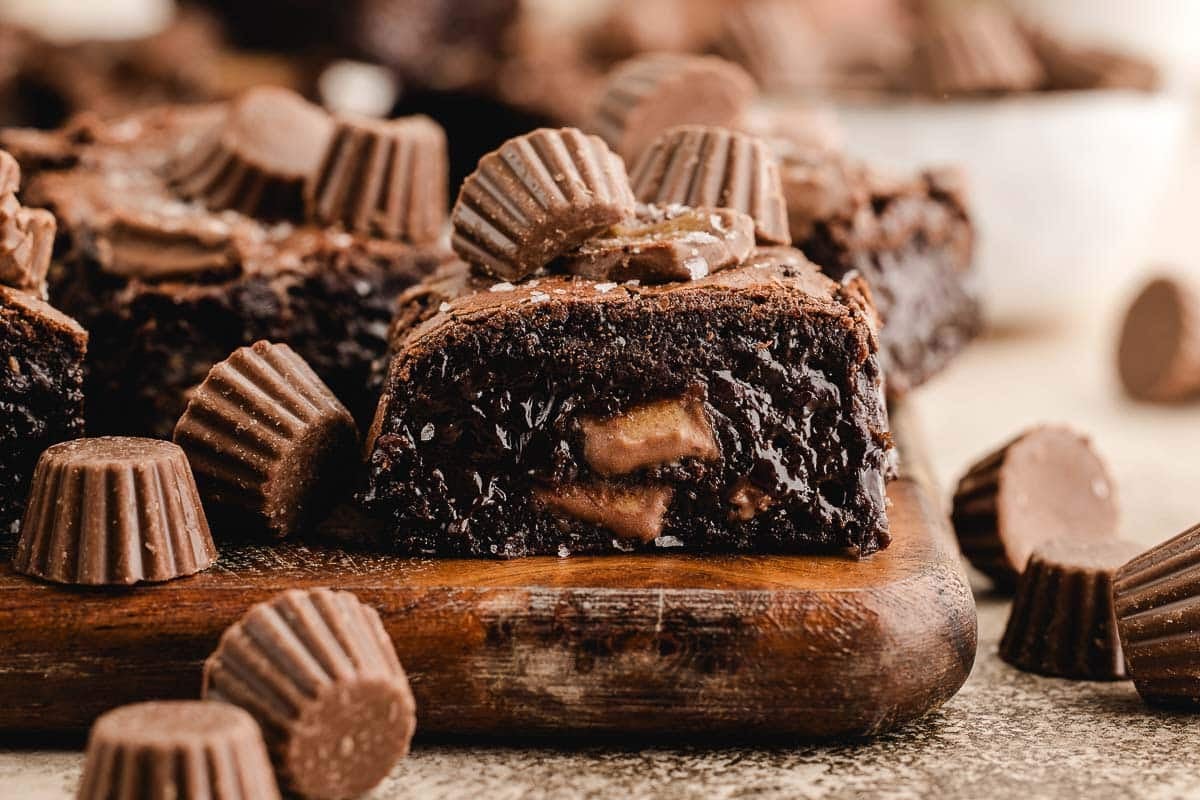 A close-up of a chocolate brownie with pieces of mini peanut butter cups both inside and on top, displayed on a wooden board.