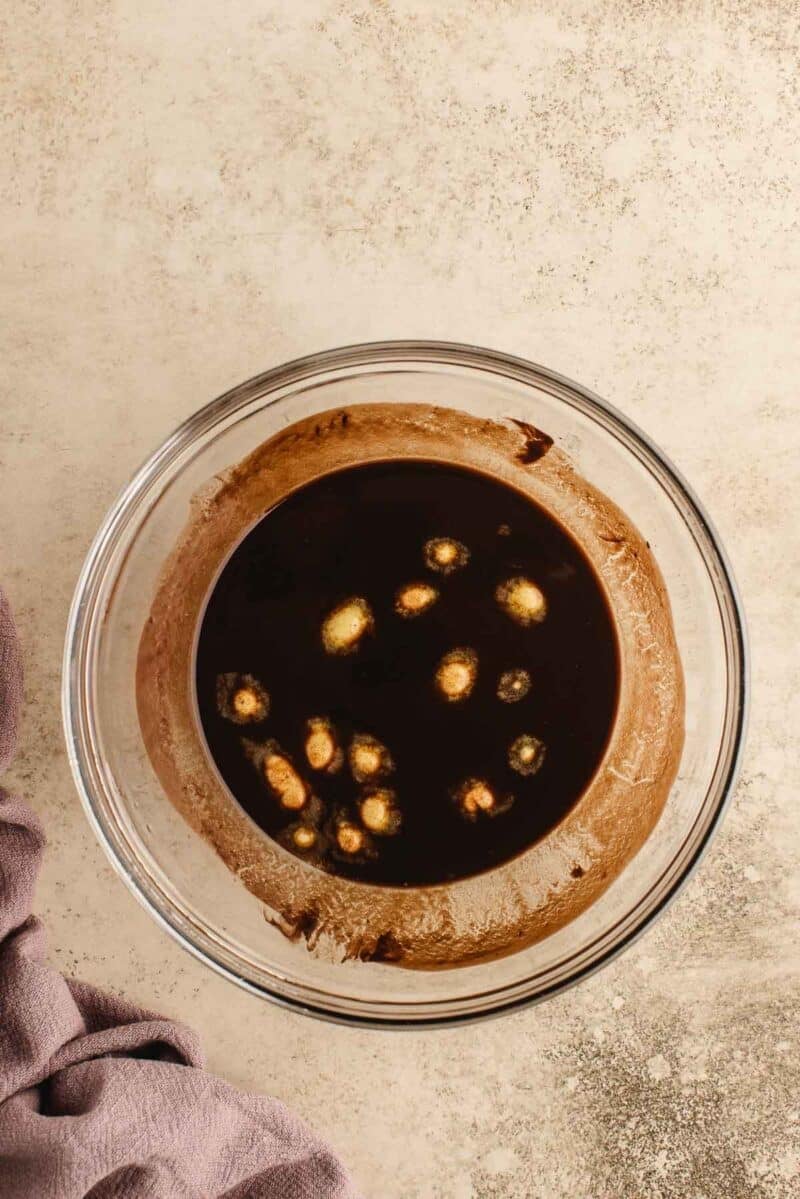 A glass mixing bowl containing chocolate brownie batter with melted butter on top, placed on a light textured surface next to a purple cloth.