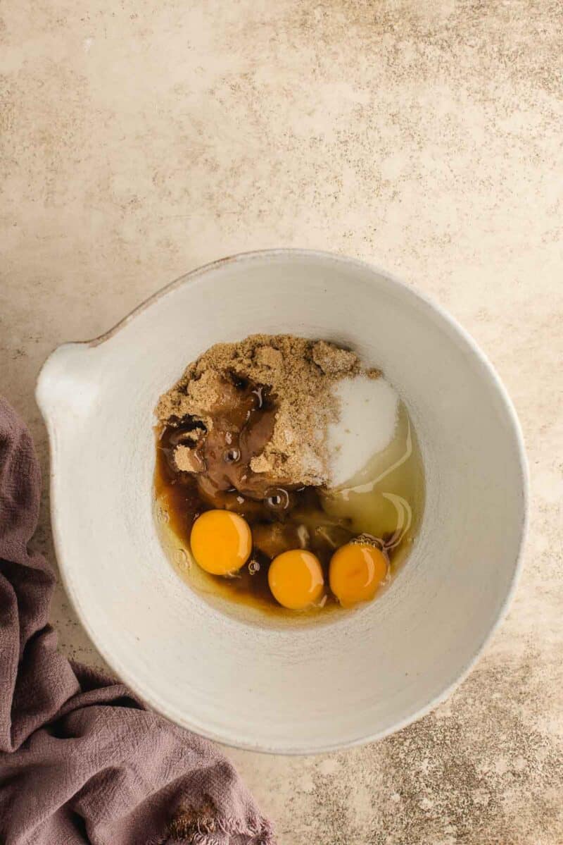 A white mixing bowl containing three eggs, brown sugar, white sugar, and vanilla extract on a beige surface next to a purple cloth.