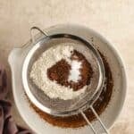 A metal sifter holds flour, cocoa powder, and salt above a mixing bowl with wet ingredients. A purple cloth is beside the bowl on a light textured surface.