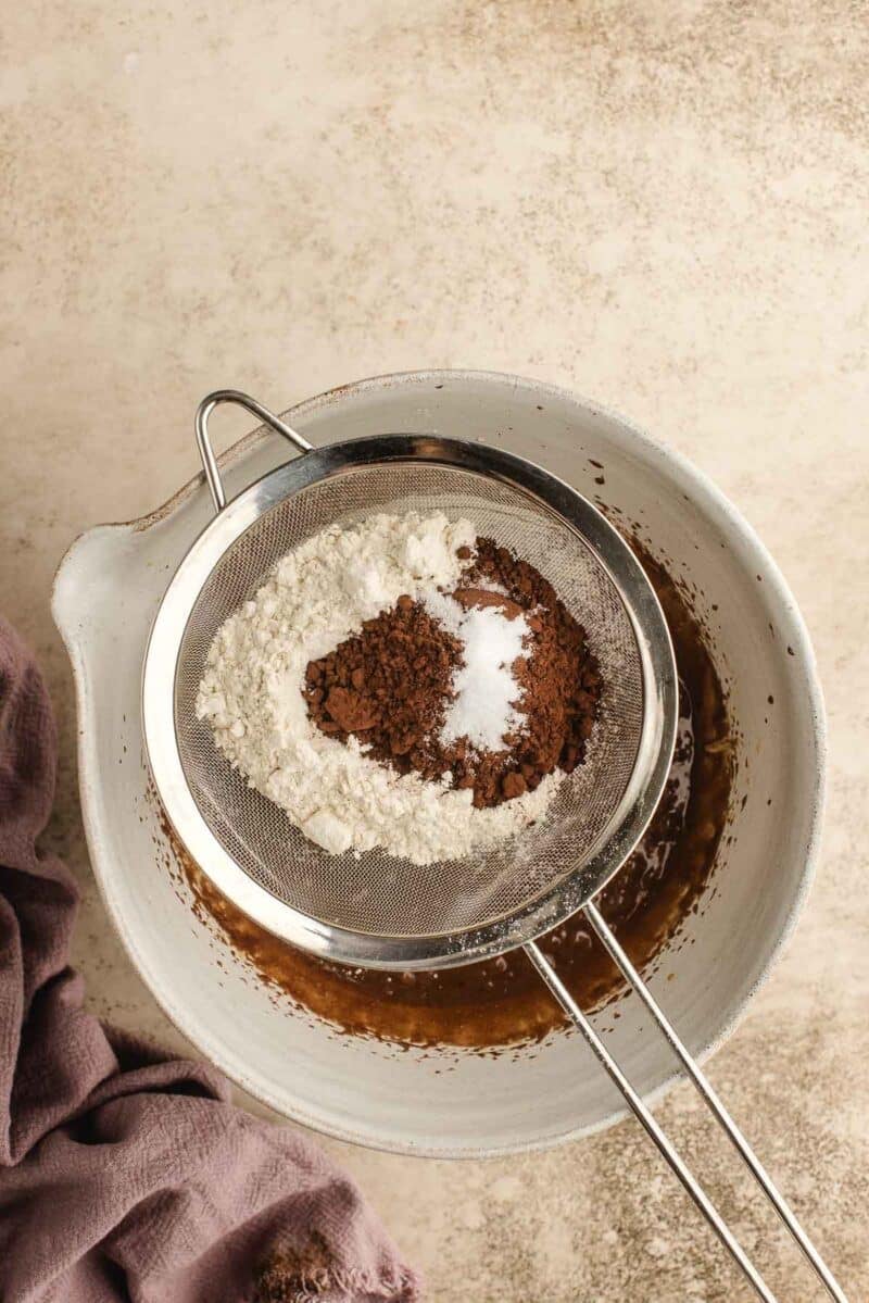 A metal sifter holds flour, cocoa powder, and salt above a mixing bowl with wet ingredients. A purple cloth is beside the bowl on a light textured surface.