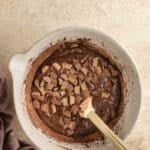 A mixing bowl containing chocolate batter with chopped peanut butter cups and a wooden spoon on a light counter.
