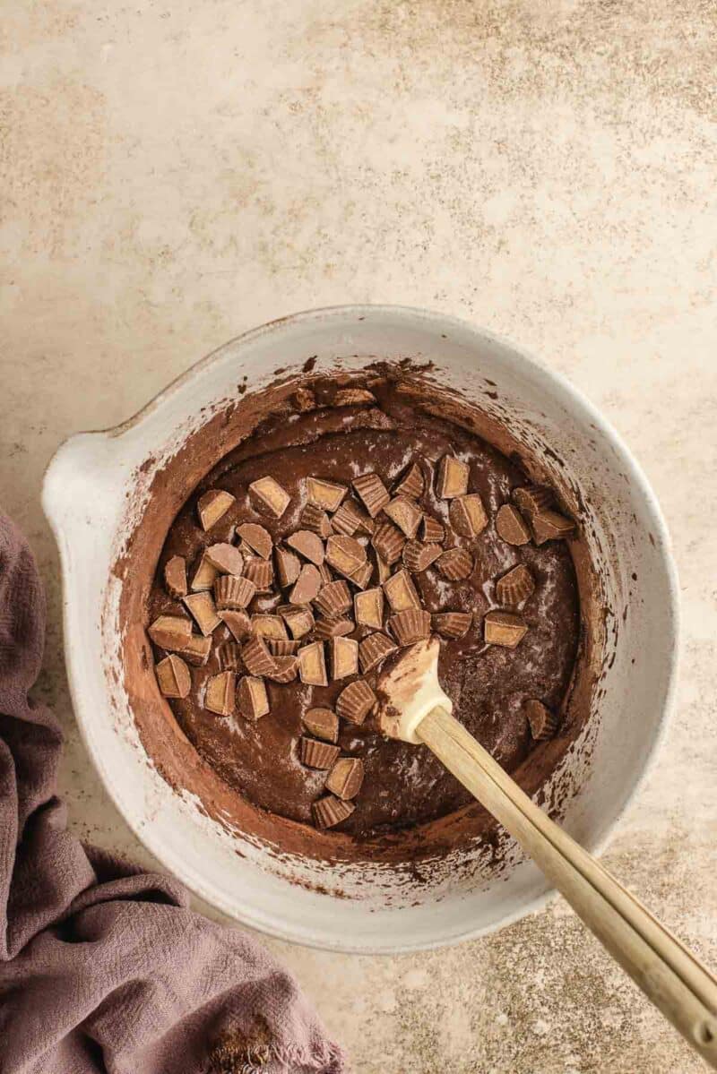 A mixing bowl containing chocolate batter with chopped peanut butter cups and a wooden spoon on a light counter.