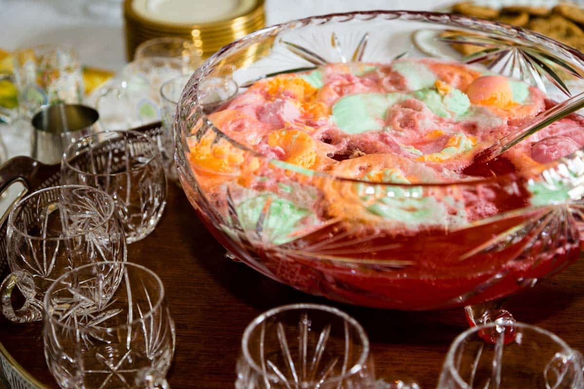 A crystal punch bowl filled with colorful sherbet punch surrounded by matching glass cups and plates on a wooden table.