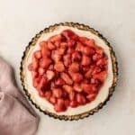 A strawberry tart with a creamy filling and a nutty crust sits on a pale background next to a pink napkin.