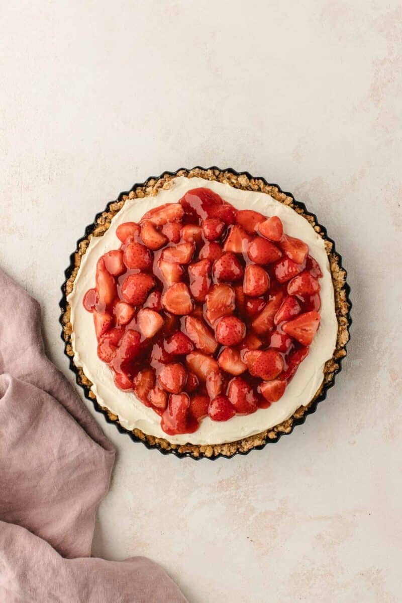 A strawberry tart with a creamy filling and a nutty crust sits on a pale background next to a pink napkin.