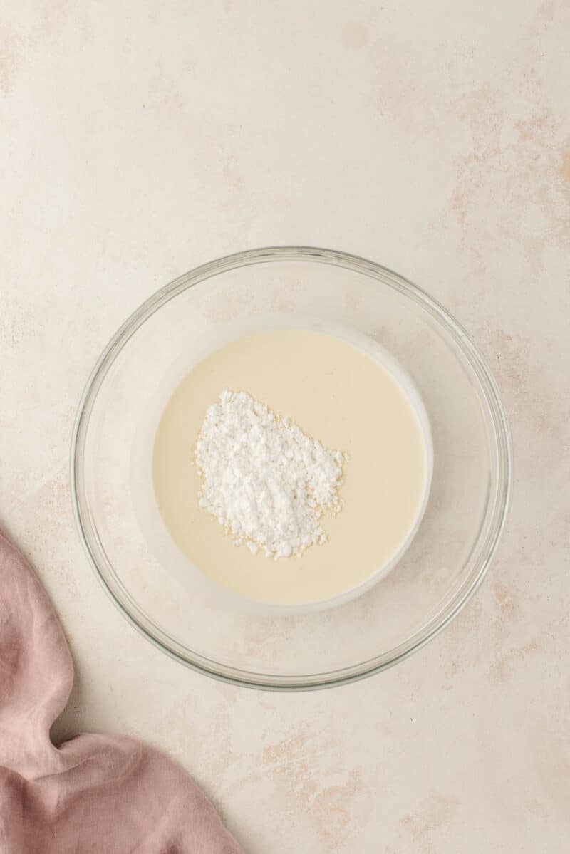A pile of powdered sugar rests on top of a light-colored liquid in a clear glass bowl.