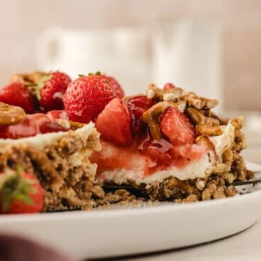 A slice of strawberry pretzel pie with fresh strawberries and pecans on a white plate, with a fork taking a bite.