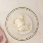 A glass bowl containing white sugar and softened butter, viewed from above, on a light surface with a pink linen nearby.