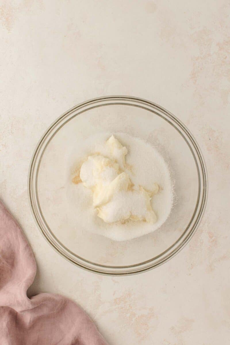 A glass bowl containing white sugar and softened butter, viewed from above, on a light surface with a pink linen nearby.