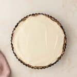 A cream-filled tart with a textured crust sits in a fluted tart pan on a light background.