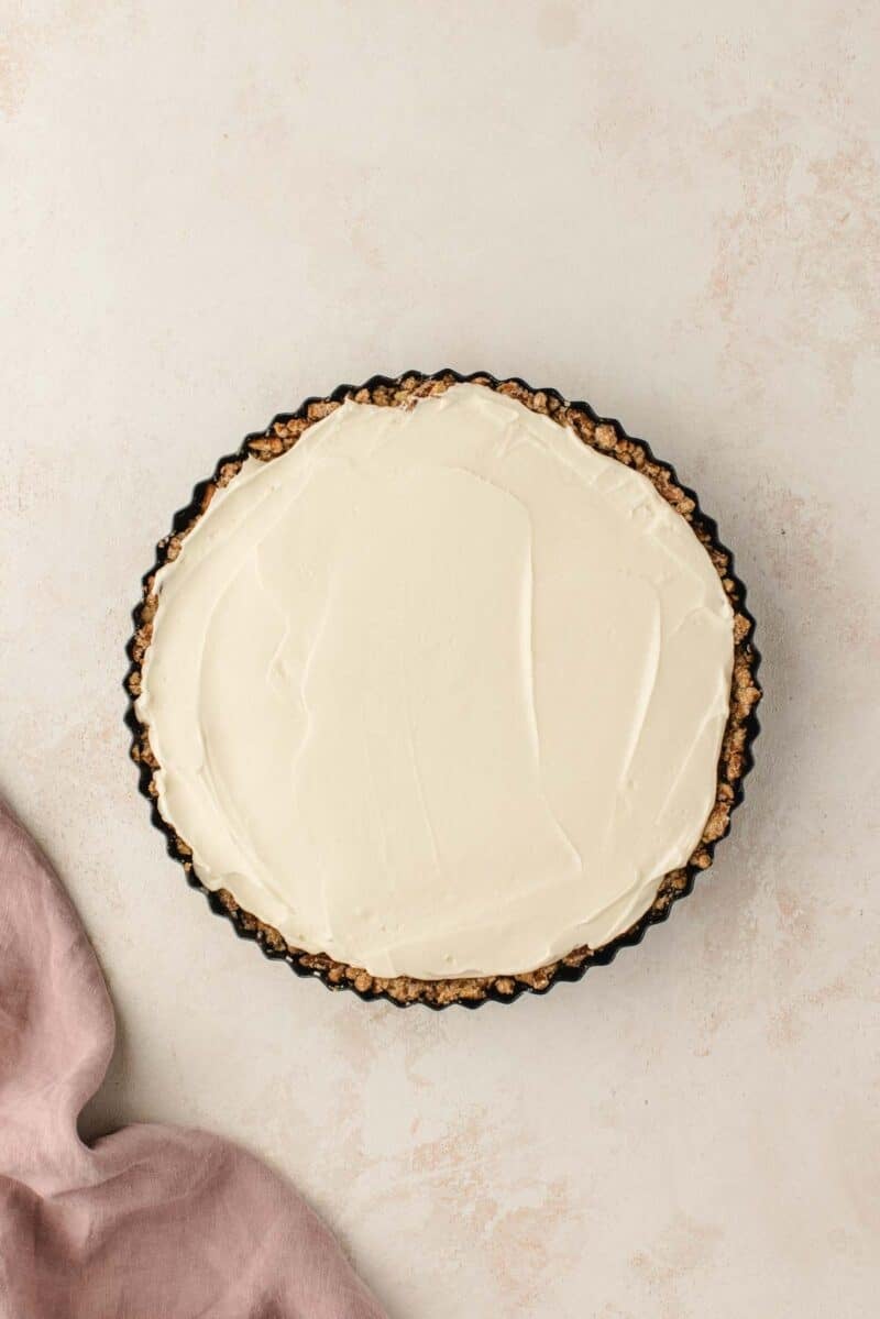 A cream-filled tart with a textured crust sits in a fluted tart pan on a light background.