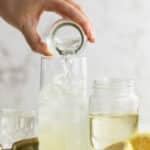 A hand pours liquid from a small glass into a tall glass filled with ice, next to a jar, a cut lemon, and a jigger on a white surface.