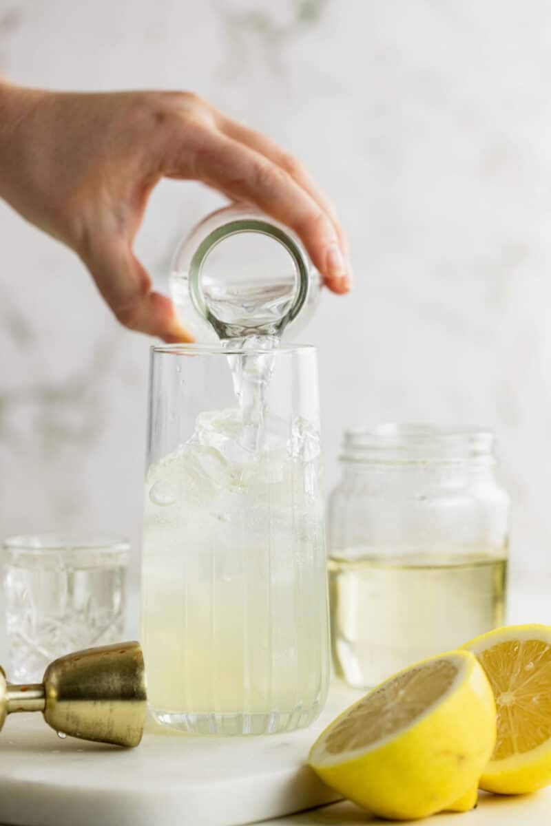 A hand pours liquid from a small glass into a tall glass filled with ice, next to a jar, a cut lemon, and a jigger on a white surface.