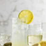 A glass of Vodka Collins with ice and a lemon slice garnish sits on a table, surrounded by lemon halves, a cocktail jigger, and glass jars.