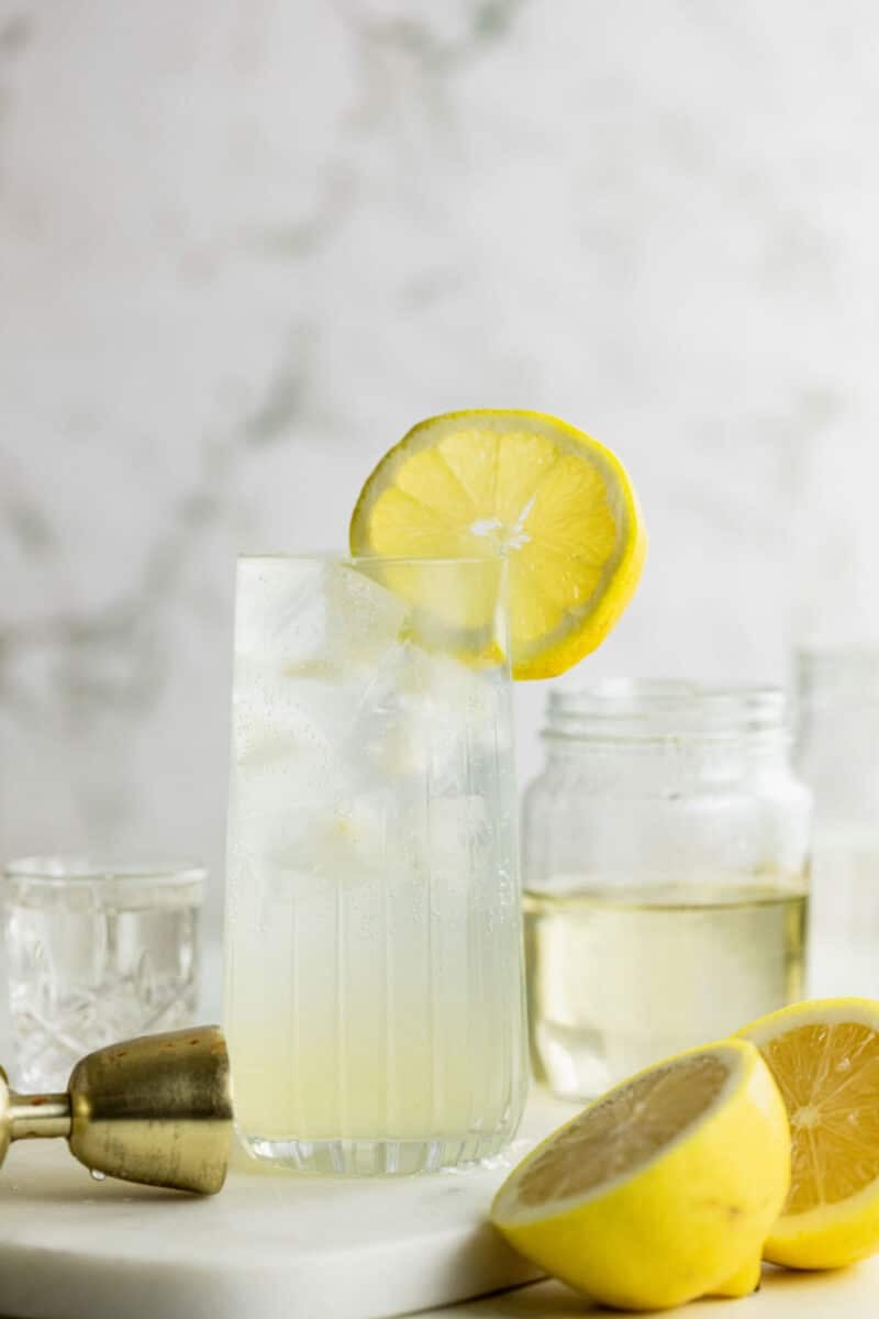 A glass of Vodka Collins with ice and a lemon slice garnish sits on a table, surrounded by lemon halves, a cocktail jigger, and glass jars.