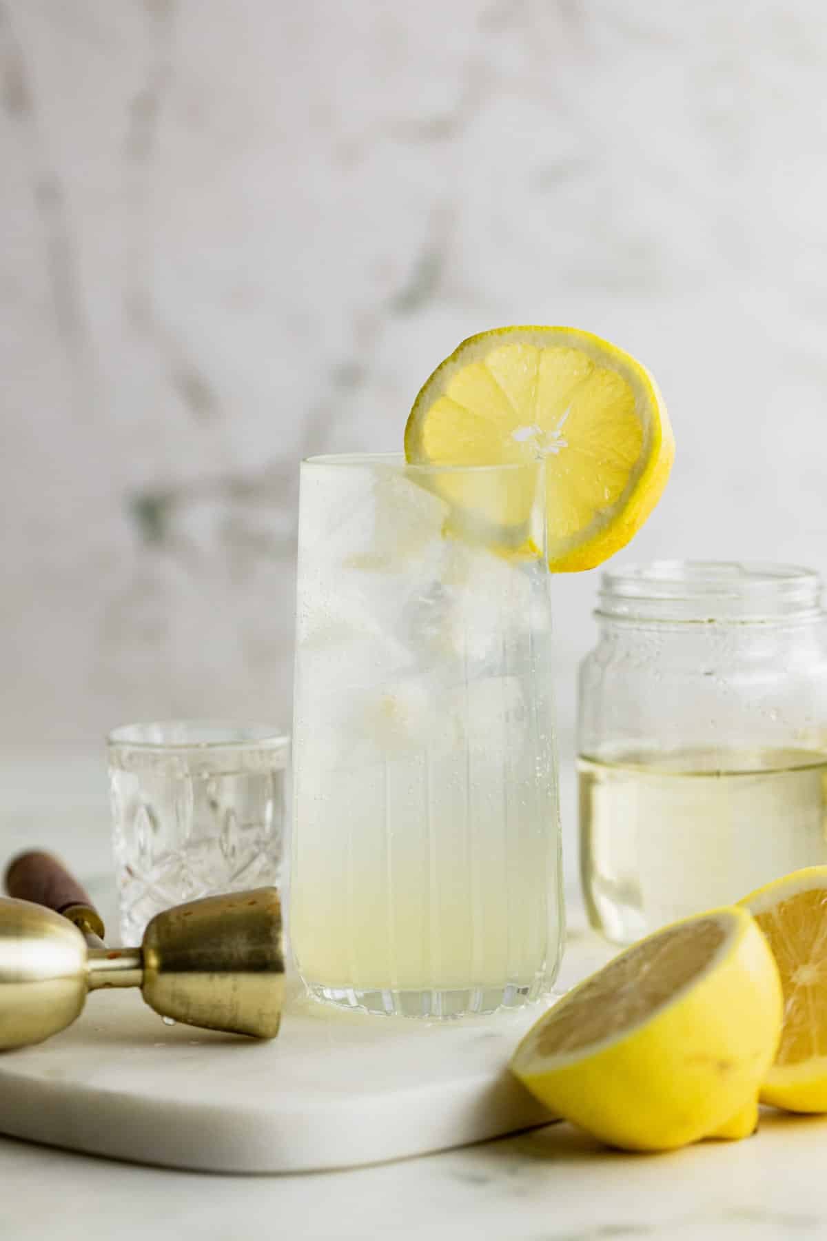 A tall glass of Vodka Collins with ice and a lemon slice garnish sits on a marble surface, alongside a halved lemon, a glass jar, and a cocktail strainer.