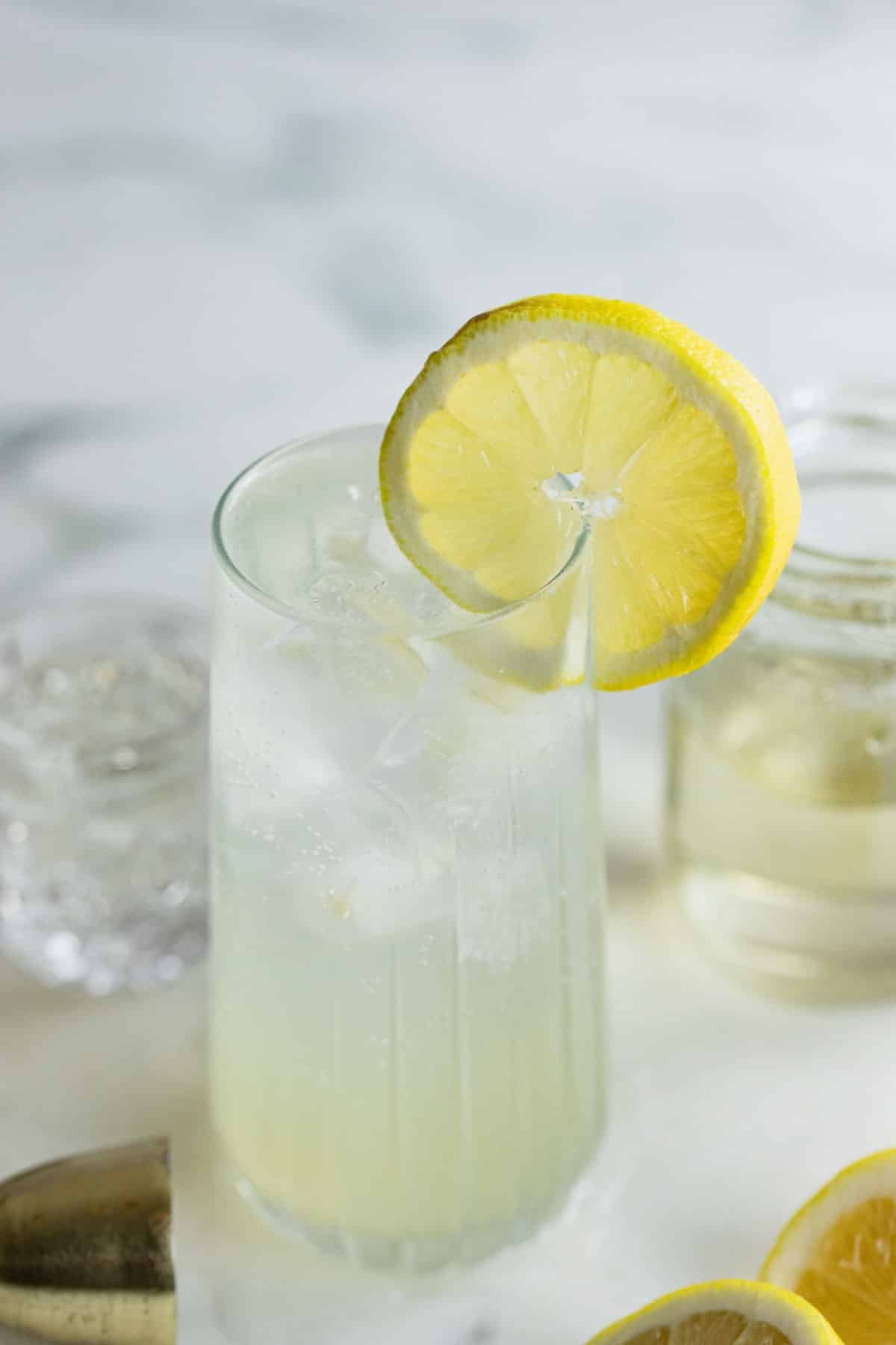 A glass of Vodka Collins with a lemon slice garnish sits on a white surface, next to a clear jar and scattered lemon slices.