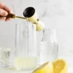 A hand pours liquid from a jigger into a tall glass containing lemon juice, with a halved lemon, a glass of water, and a water jar nearby.
