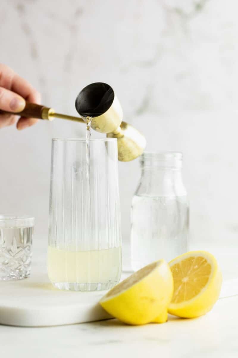 A hand pours liquid from a jigger into a tall glass containing lemon juice, with a halved lemon, a glass of water, and a water jar nearby.
