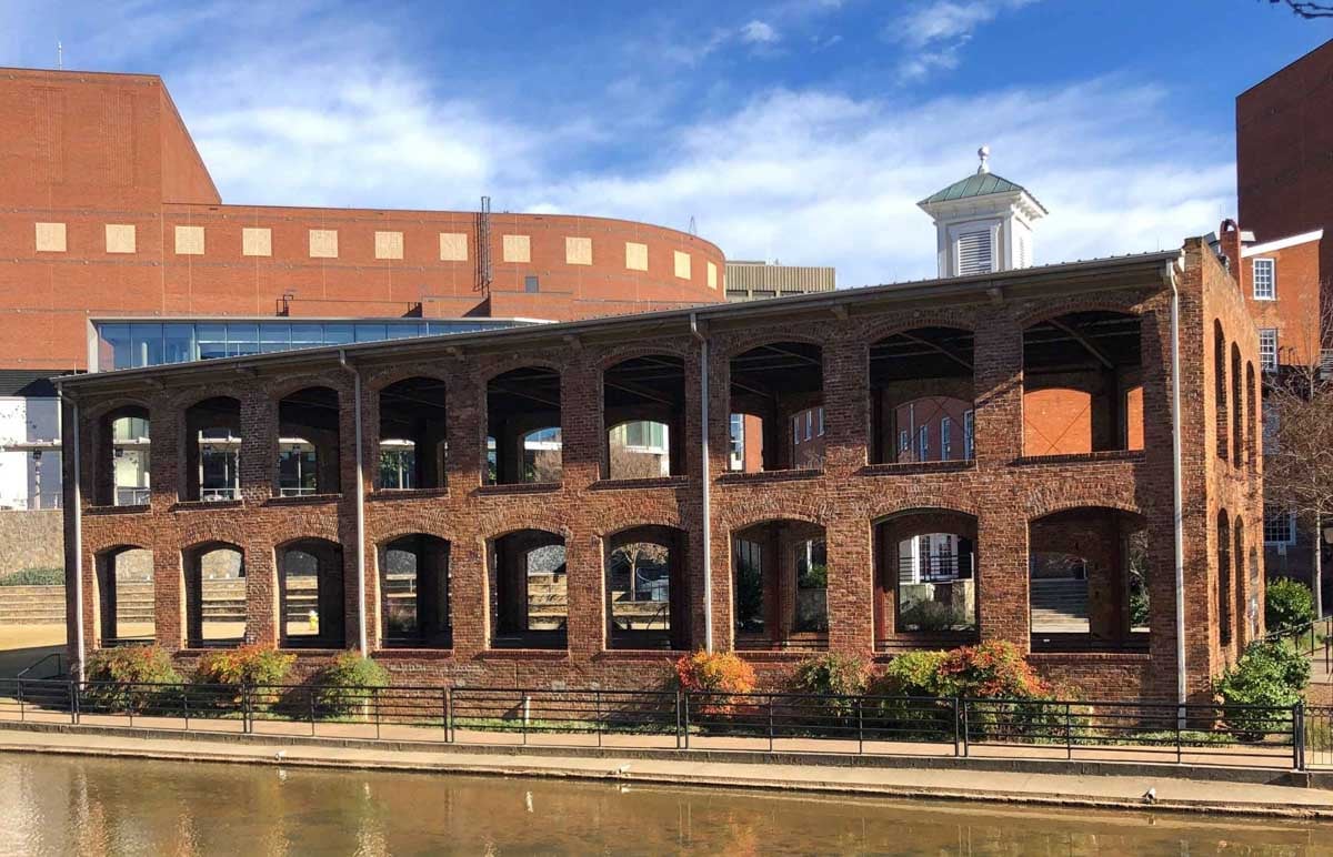 A brick building with open archways and no windows stands beside a canal, with a clear blue sky in the background.