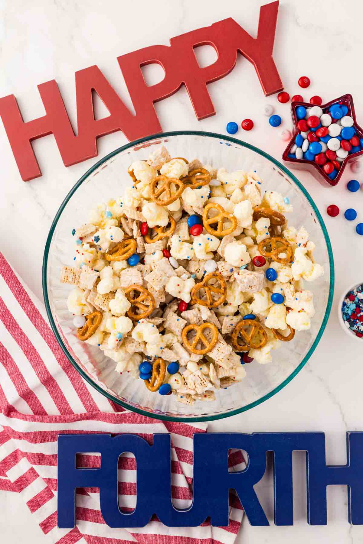 A bowl of snack mix with popcorn, pretzels, cereal, and red, white, and blue candies, surrounded by “HAPPY FOURTH” letters and festive decorations.