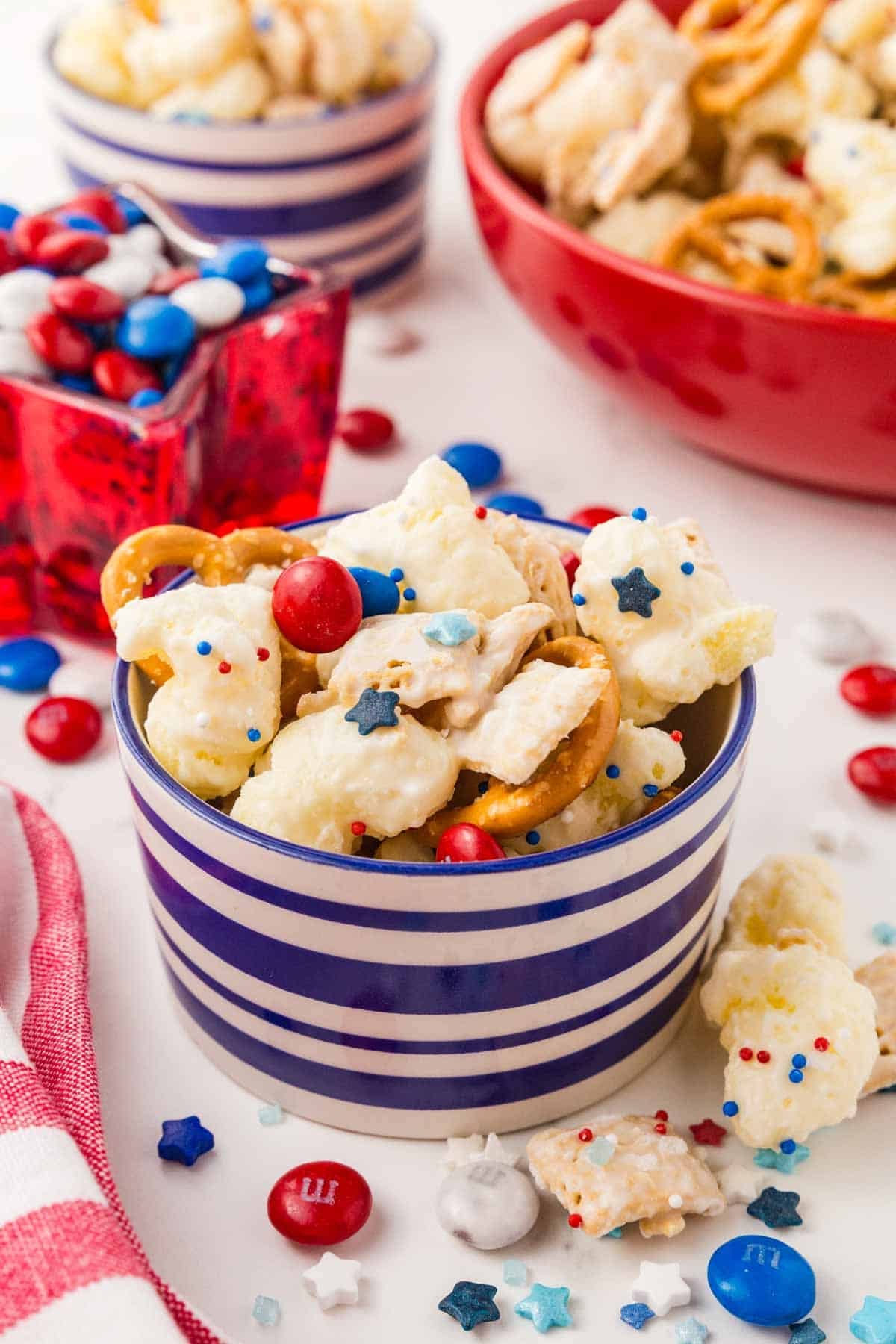 A striped bowl filled with snack mix, including pretzels, popcorn, and red, white, and blue candies, sits on a table with more candies and snacks in the background.