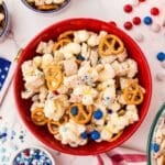 A red bowl filled with popcorn, pretzels, cereal, mini marshmallows, and red, white, and blue candies, surrounded by patriotic decorations and snacks.