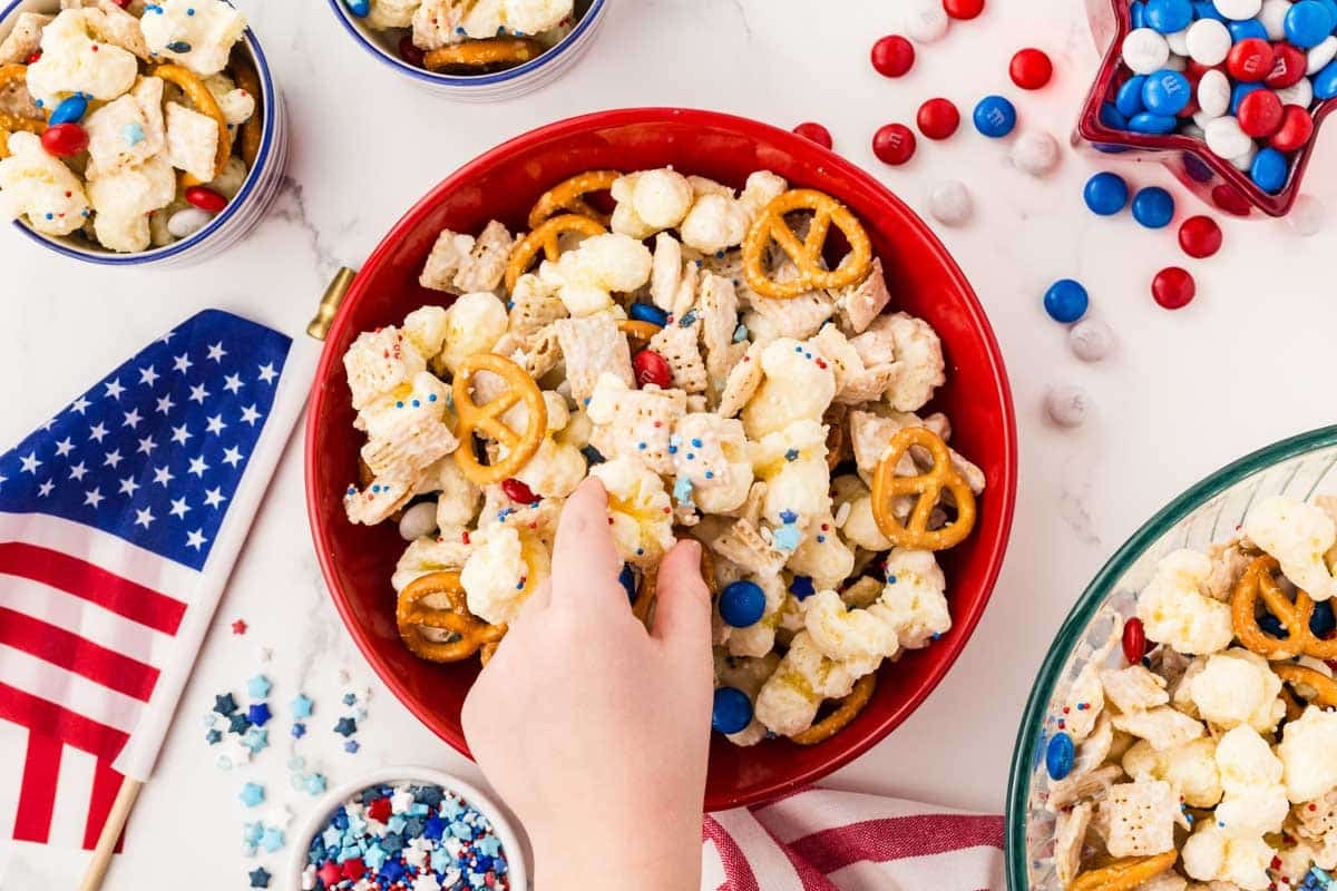A hand reaches into a red bowl filled with a snack mix of pretzels, popcorn, cereal, and red, white, and blue candies. An American flag and festive decorations are nearby.