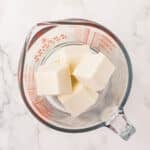 Glass measuring cup with several rectangular blocks of white butter or shortening on a white marble surface.