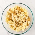 A glass bowl filled with popcorn and square cereal pieces on a white marble surface.