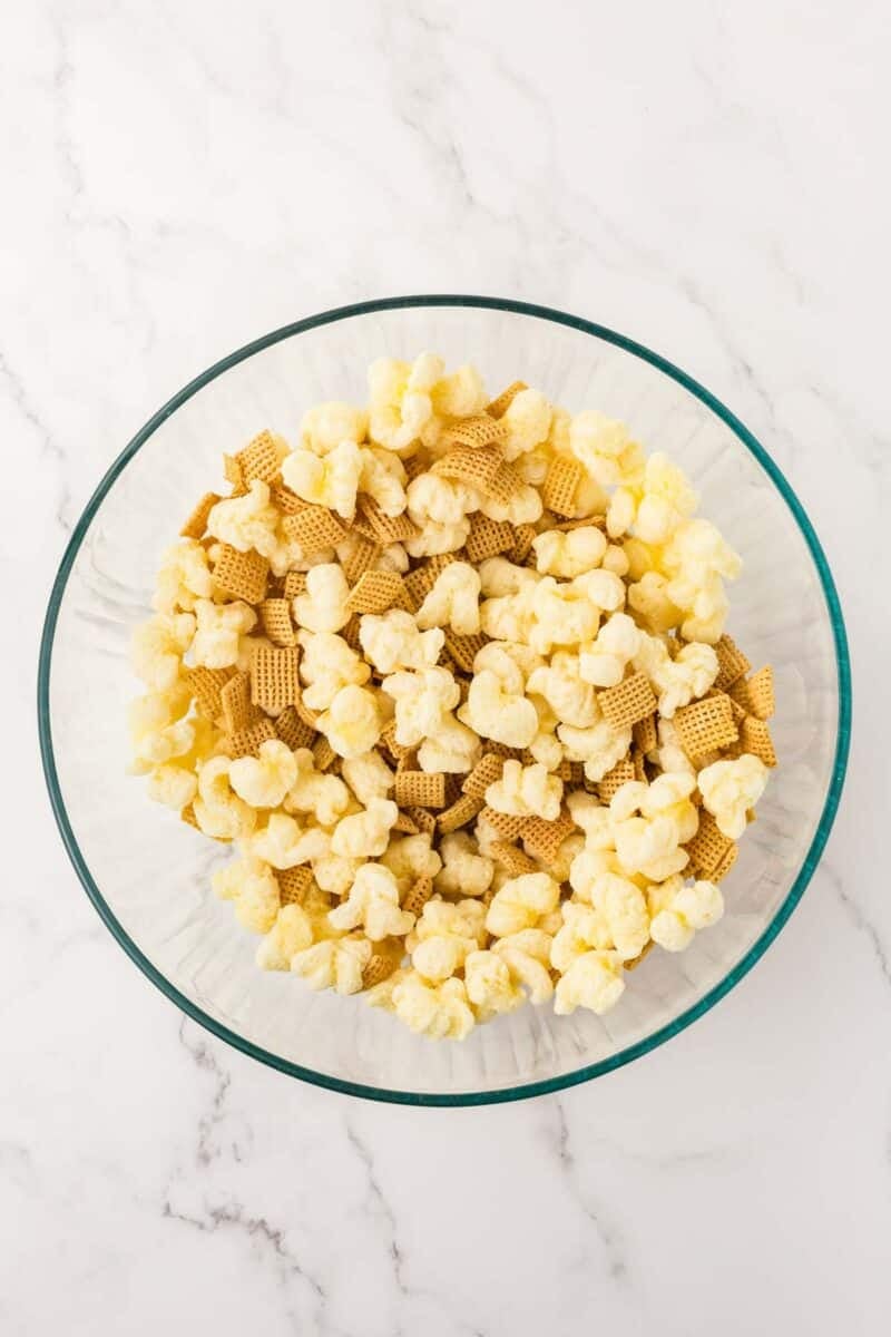 A glass bowl filled with popcorn and square cereal pieces on a white marble surface.