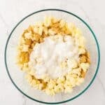 A glass bowl containing popcorn, Chex cereal, and a layer of white melted coating on top, placed on a marble surface.