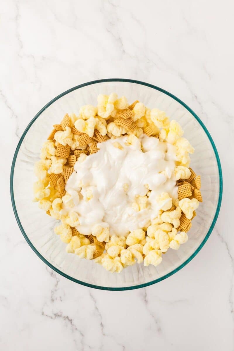 A glass bowl containing popcorn, Chex cereal, and a layer of white melted coating on top, placed on a marble surface.