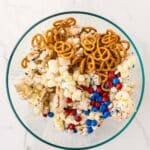 A glass bowl filled with pretzels, popcorn, Chex cereal, and red, white, and blue chocolate candies on a white marble surface.