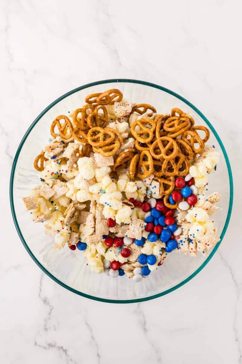 A glass bowl filled with pretzels, popcorn, Chex cereal, and red, white, and blue chocolate candies on a white marble surface.