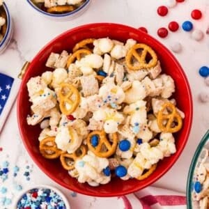 A red bowl filled with a snack mix of pretzels, popcorn, cereal pieces, and red, white, and blue candy-coated chocolates, on a white surface with scattered candies and sprinkles nearby.