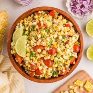 A wooden bowl filled with corn salad, cherry tomatoes, red onion, cilantro, and lime wedges. Surrounding the bowl are lime halves, chopped onion, and corn kernels.
