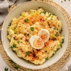A bowl of potato salad garnished with paprika, parsley, and sliced boiled eggs, placed on a woven mat with a fork nearby.