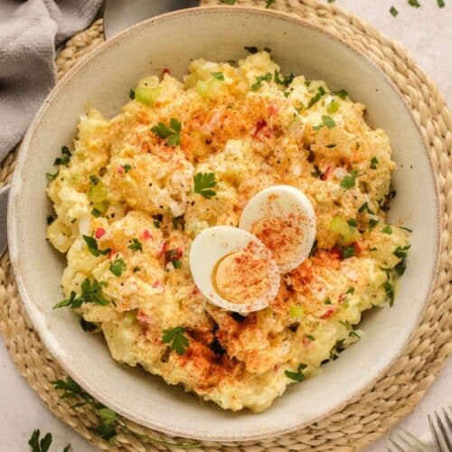A bowl of potato salad garnished with paprika, parsley, and sliced boiled eggs, placed on a woven mat with a fork nearby.
