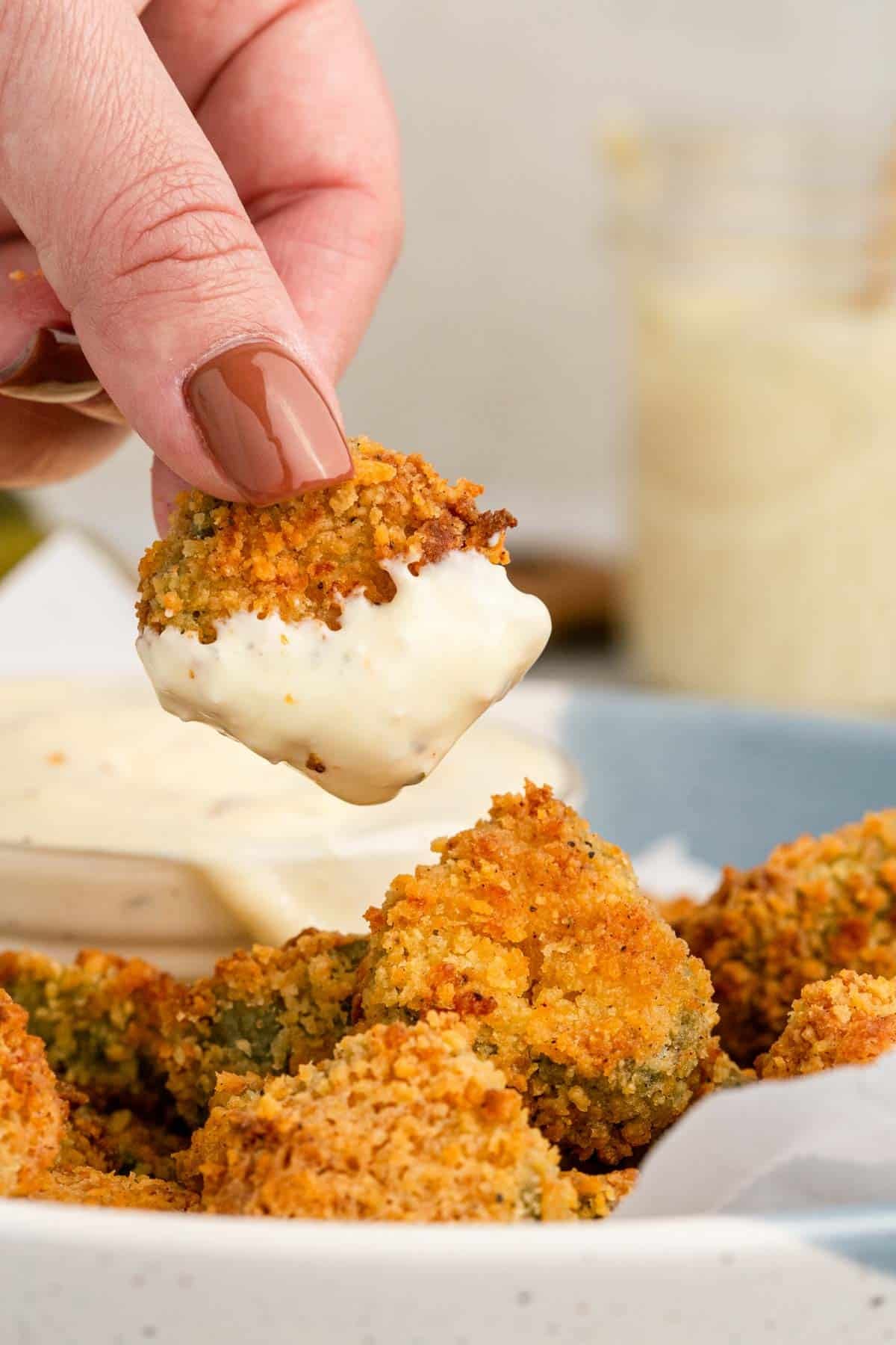 A hand holding a breaded and fried pickle chip dipped in white sauce, with more fried pickle chips on a plate below.