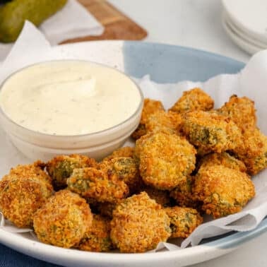 A plate of air fryer fried pickles is served with a bowl of white dipping sauce on parchment paper.