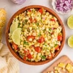 A wooden bowl of corn salad with cherry tomatoes, red onion, cilantro, and lime wedges, surrounded by fresh corn, lime halves, and chopped ingredients on a light surface.