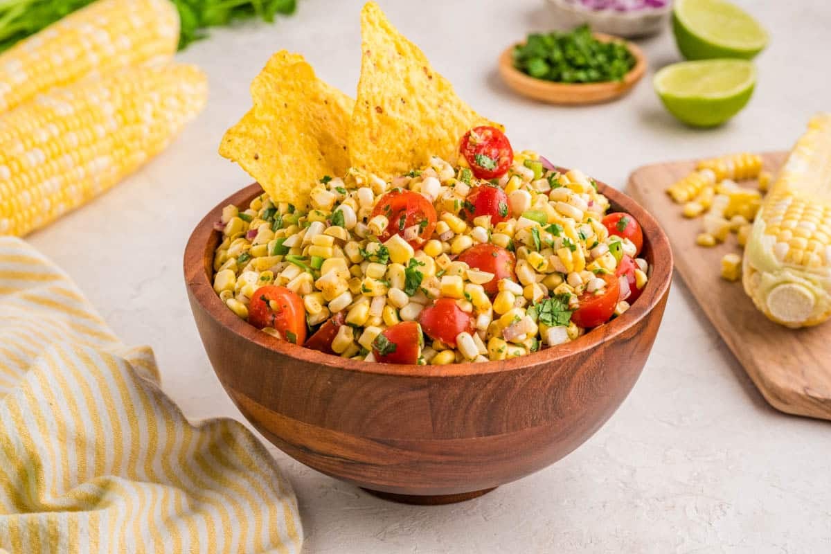 A wooden bowl filled with corn salad, cherry tomatoes, herbs, and two tortilla chips, with corn cobs, lime halves, and chopped vegetables in the background.