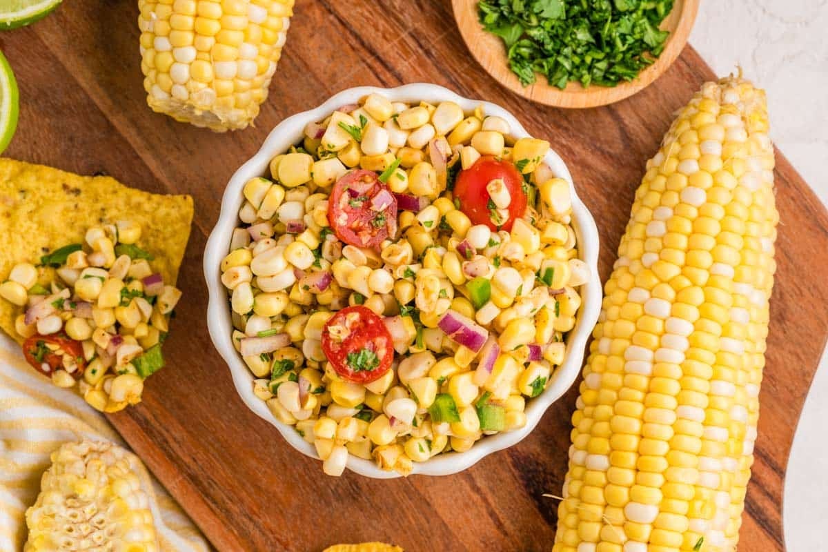A bowl of corn salad with cherry tomatoes, red onion, and herbs on a wooden board, surrounded by fresh corn, chopped parsley, and lime wedges.