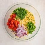 A glass bowl containing chopped cilantro, corn, cherry tomatoes, red onion, and jalapeño, arranged in separate sections on a light surface.
