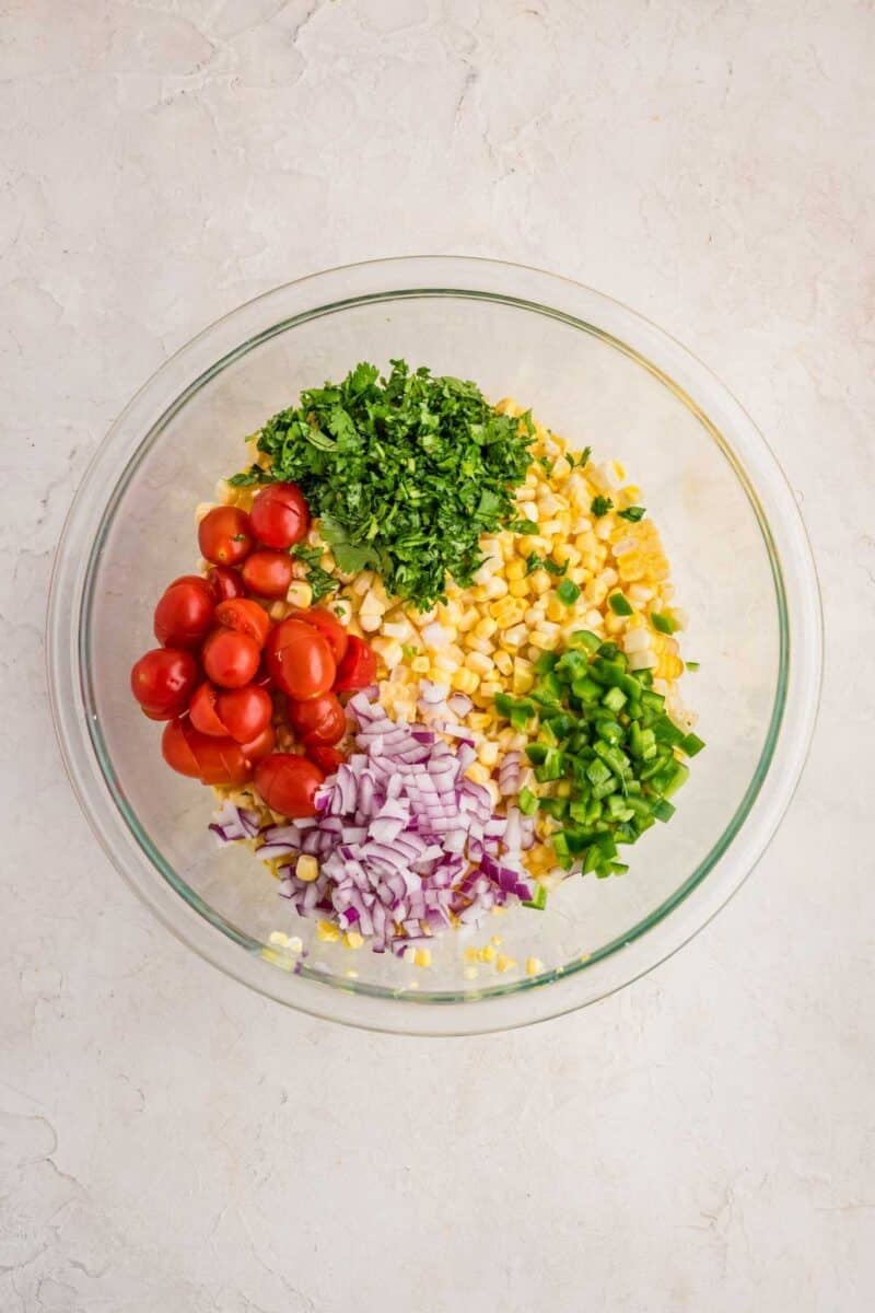 A glass bowl containing chopped cilantro, corn, cherry tomatoes, red onion, and jalapeño, arranged in separate sections on a light surface.