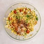 A glass bowl containing a mixture of corn, chopped tomatoes, red onion, cilantro, and various spices on a light-colored surface.