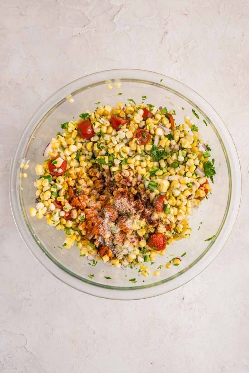 A glass bowl containing a mixture of corn, chopped tomatoes, red onion, cilantro, and various spices on a light-colored surface.